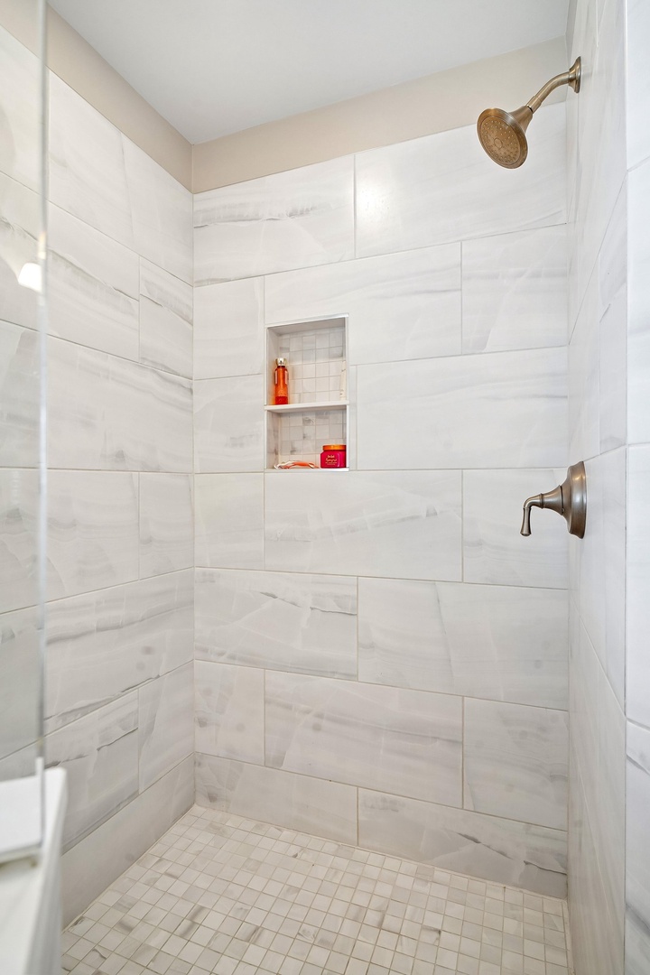 Step into your private spa-like sanctuary with crisp white marble-style tile, a sleek brushed-gold rainfall showerhead, and built-in shelving for all your essentials.