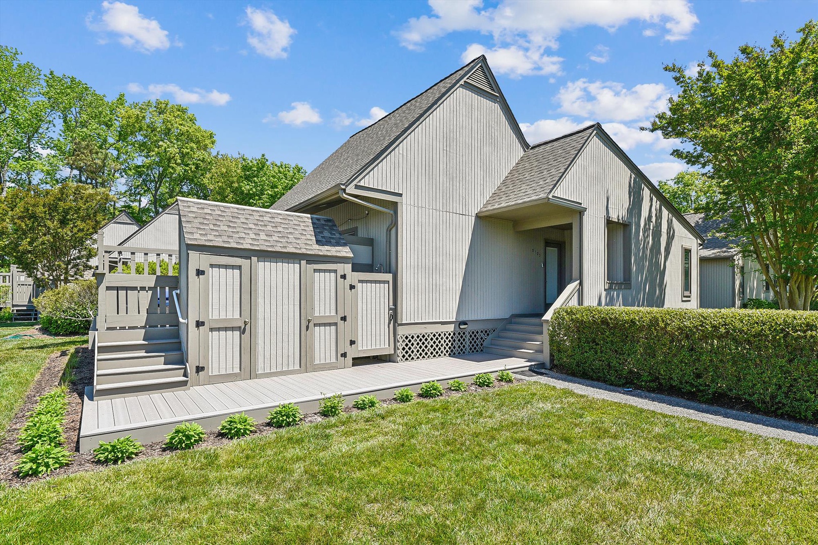 A charming white barn-style cottage nestled in a peaceful neighborhood, featuring a private shed, lush lawn, and surrounded by mature trees.