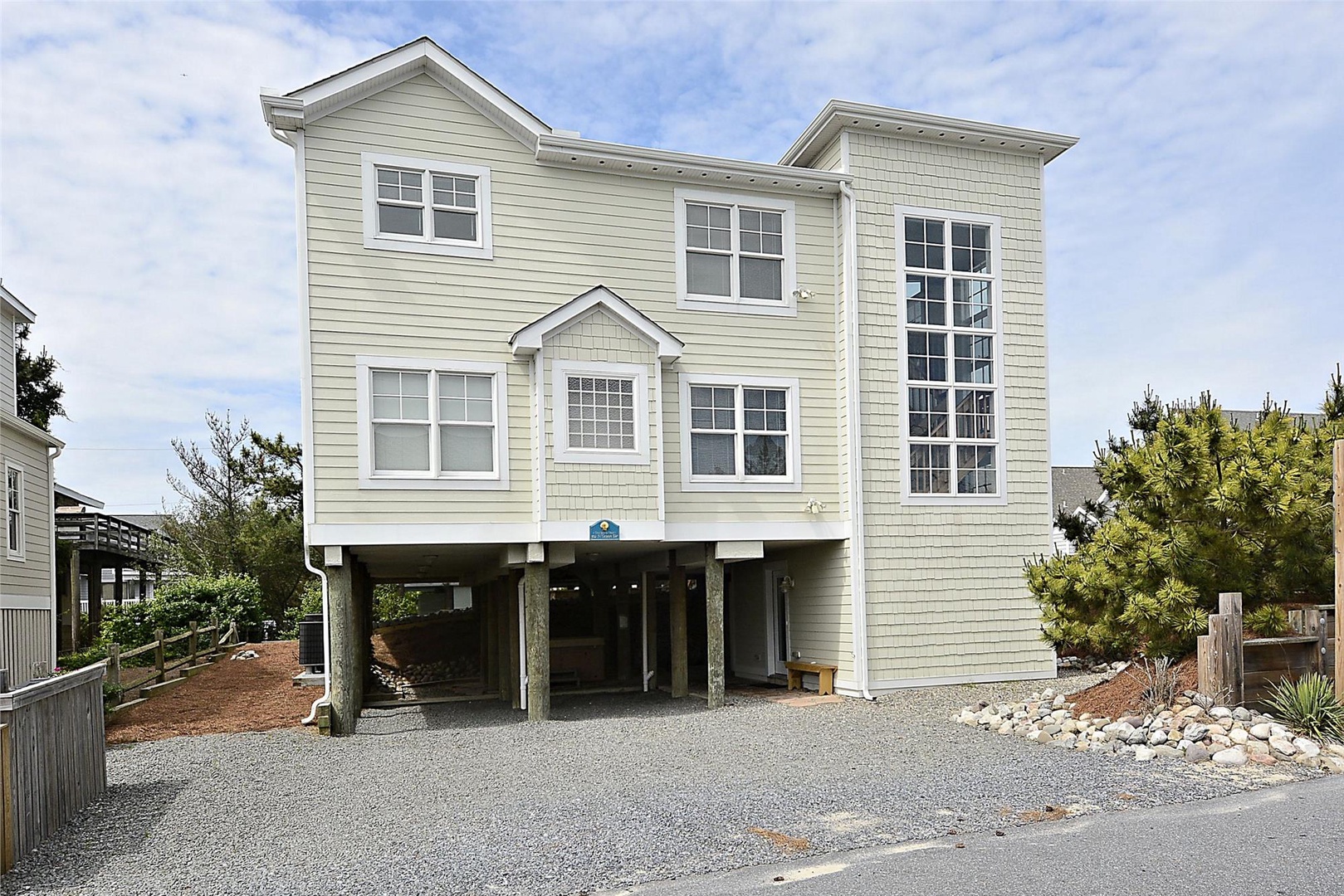 This coastal beach house features classic cottage architecture on stilts, with covered ground-level parking and mature landscaping welcoming guests to the surrounding neighborhood.