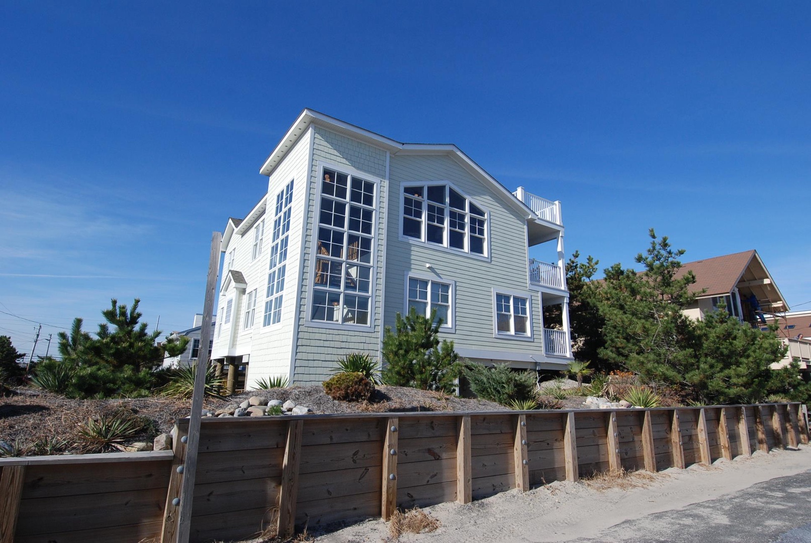 A coastal vacation home featuring expansive windows and outdoor decking, positioned in a landscaped beachside neighborhood beneath clear blue skies.