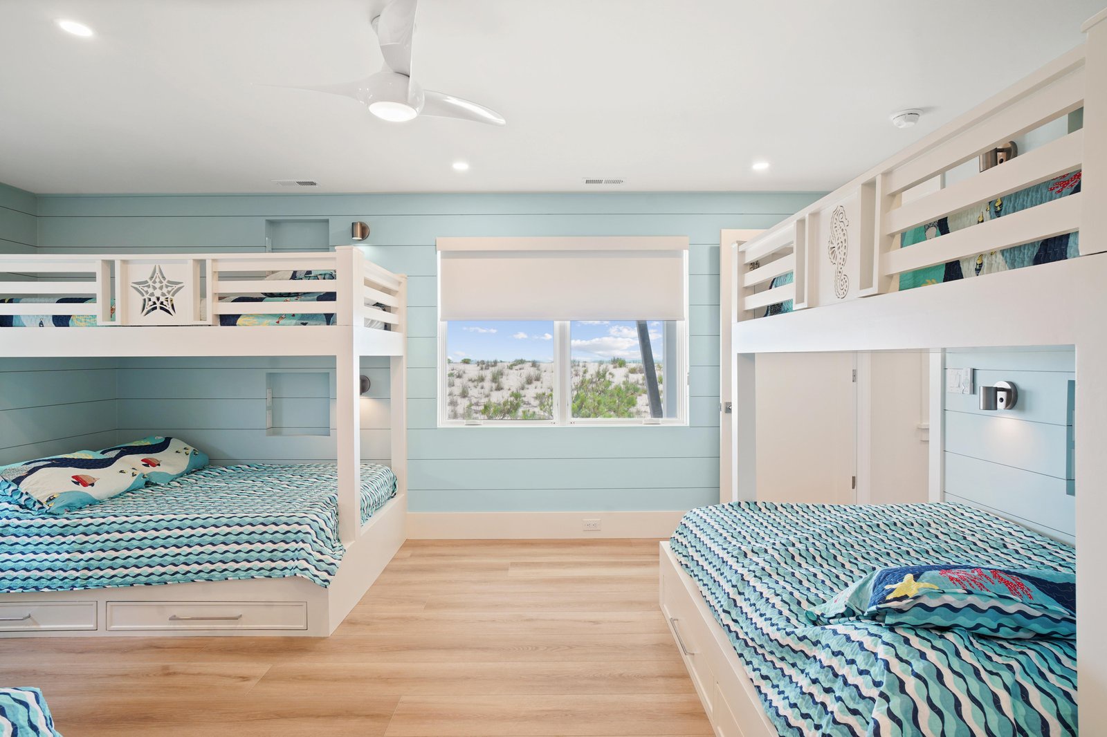 Dream of sandy toes and salty air in your cozy coastal bunk room. Each bed is nestled beneath soothing aqua walls, and a window frames gentle dunes—your perfect backdrop for a restful beach getaway.