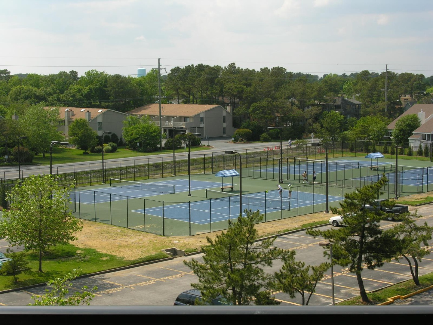 Elevated daytime view of the property's tennis courts and surrounding residential neighborhood, showcasing the recreational facilities and tree-lined area.