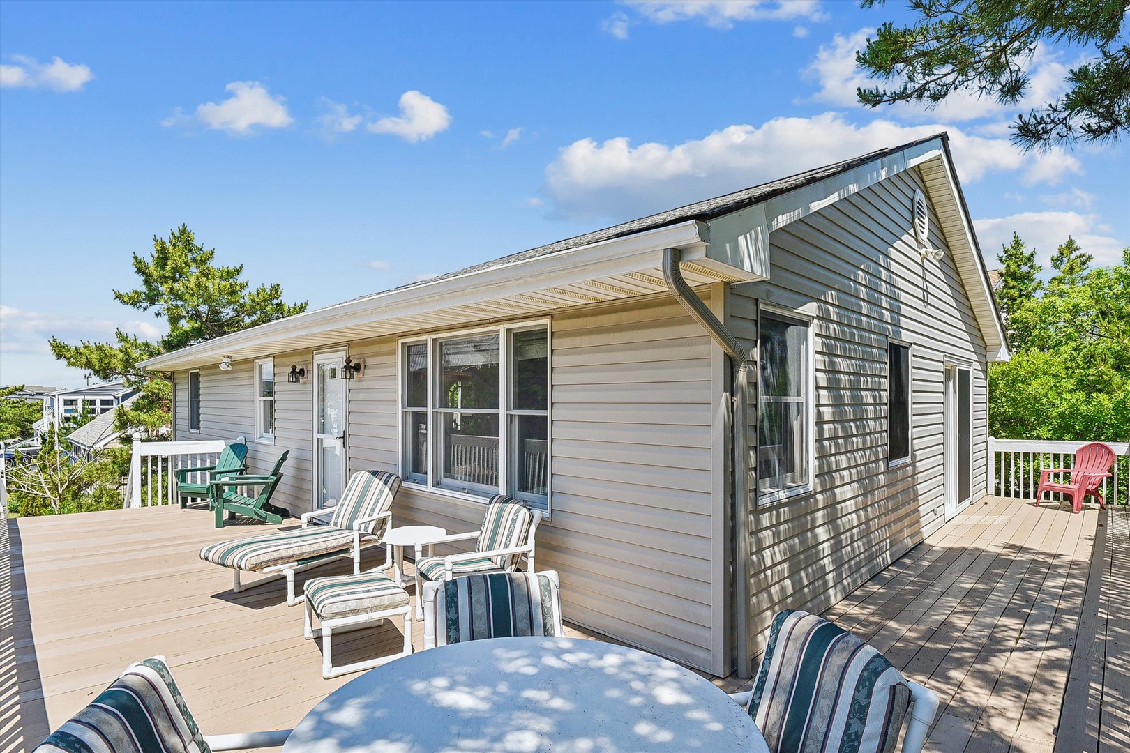 A bright sunny day frames this charming vacation home exterior with its expansive wraparound deck. The spacious outdoor living area features striped lounge chairs perfect for soaking up sunshine, green seating for morning coffee, and plenty of room for ev