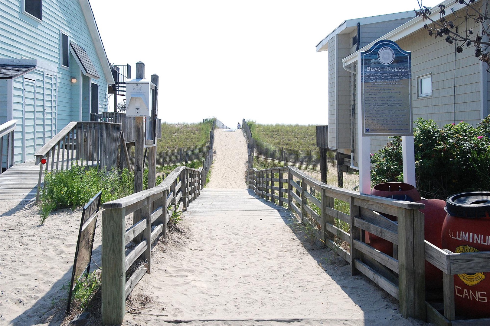 Sandy beach path flanked by dune grass and weathered wooden fencing leads from the property to the oceanfront, with beach rules posted near the access point.