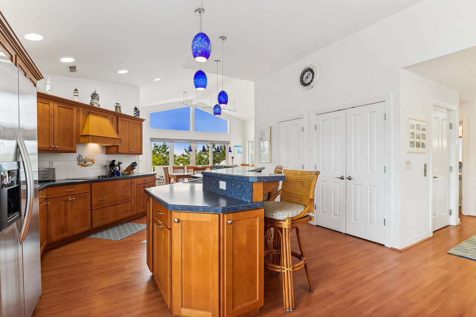 Step into your gourmet kitchen, where blue pendant lights illuminate the spacious island—perfect for morning coffee or meal prep. Warm wood cabinetry and stainless appliances invite effortless coastal cooking with garden views.