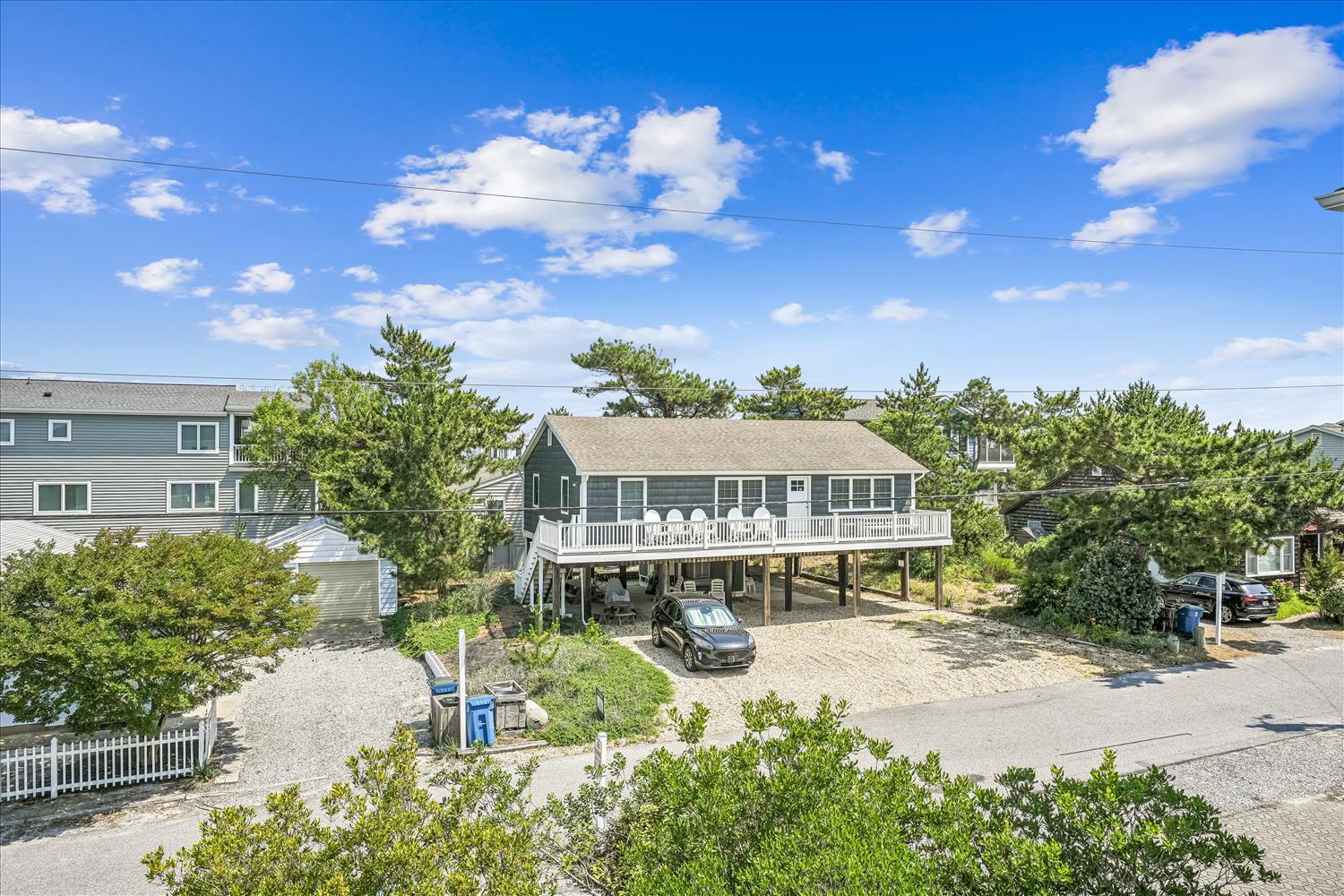 Elevated coastal beach house nestled among mature trees in a quiet residential neighborhood, offering convenient parking beneath the property and easy access to the shoreline.