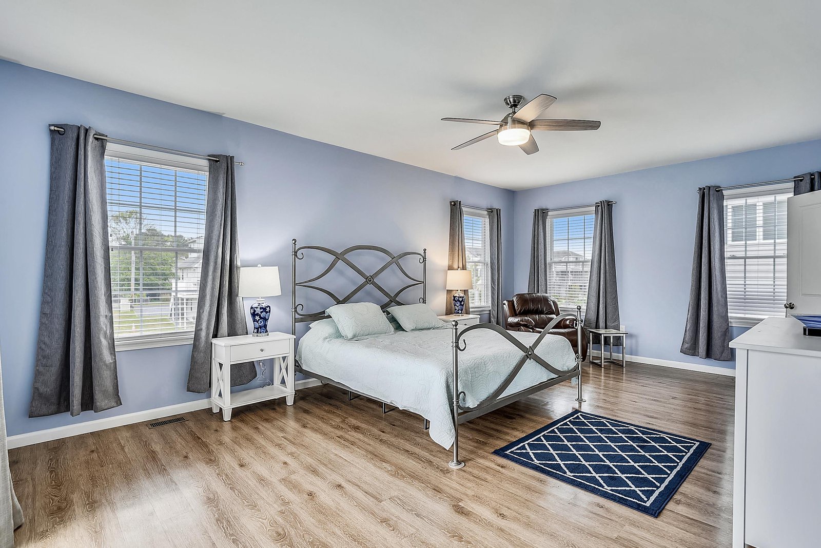 Your spacious bedroom features calming blue walls, warm wood floors, and plenty of natural light from multiple windows. Relax in the cozy reading chair or enjoy the ceiling fan overhead.