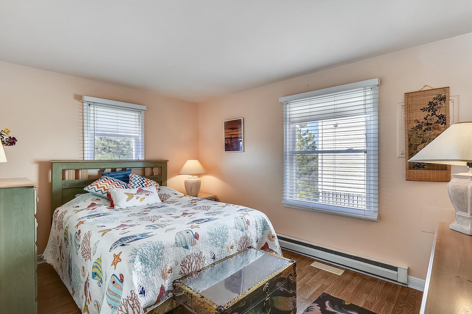 Unwind in your peaceful bedroom, where coastal-themed bedding and natural light create a serene retreat. Two windows frame leafy views, while warm tones and cozy decor invite restful nights.
