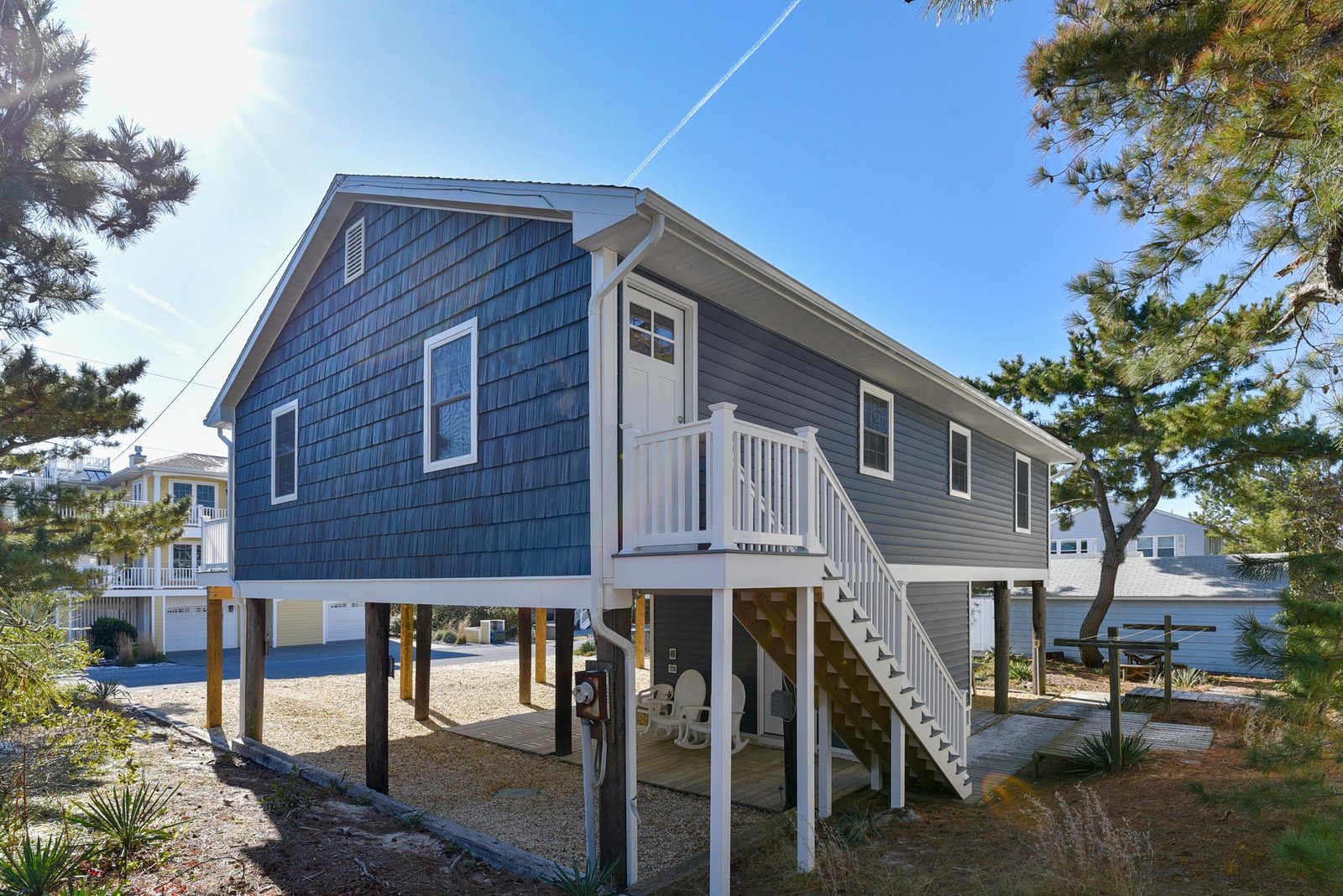 A charming coastal cottage elevated on stilts, surrounded by native landscaping and just steps from waterfront living. The covered ground-level area offers shaded parking and outdoor seating space.