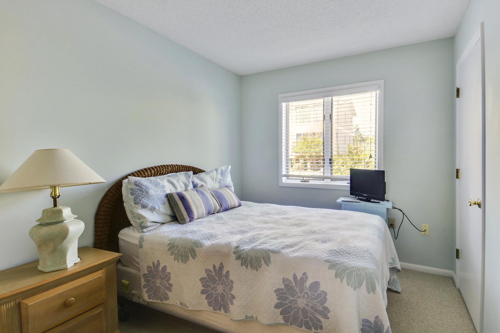 Unwind in your peaceful bedroom with soft blue walls and natural light streaming through blinds. Plush bedding and a bedside lamp create a cozy retreat for restful nights.