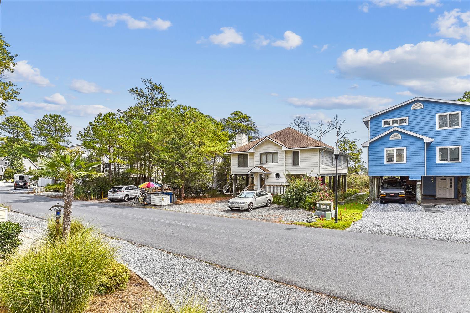 A quiet, tree-lined residential street features the distinctive blue beach house with covered parking and coastal charm—your vacation retreat awaits in this peaceful neighborhood.
