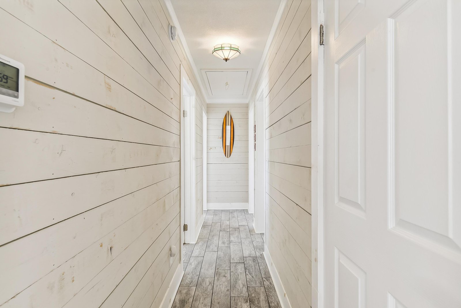 A bright hallway with white shiplap walls and weathered wood floors leads you through the home, where a surfboard accent hints at the coastal lifestyle waiting beyond these doors.