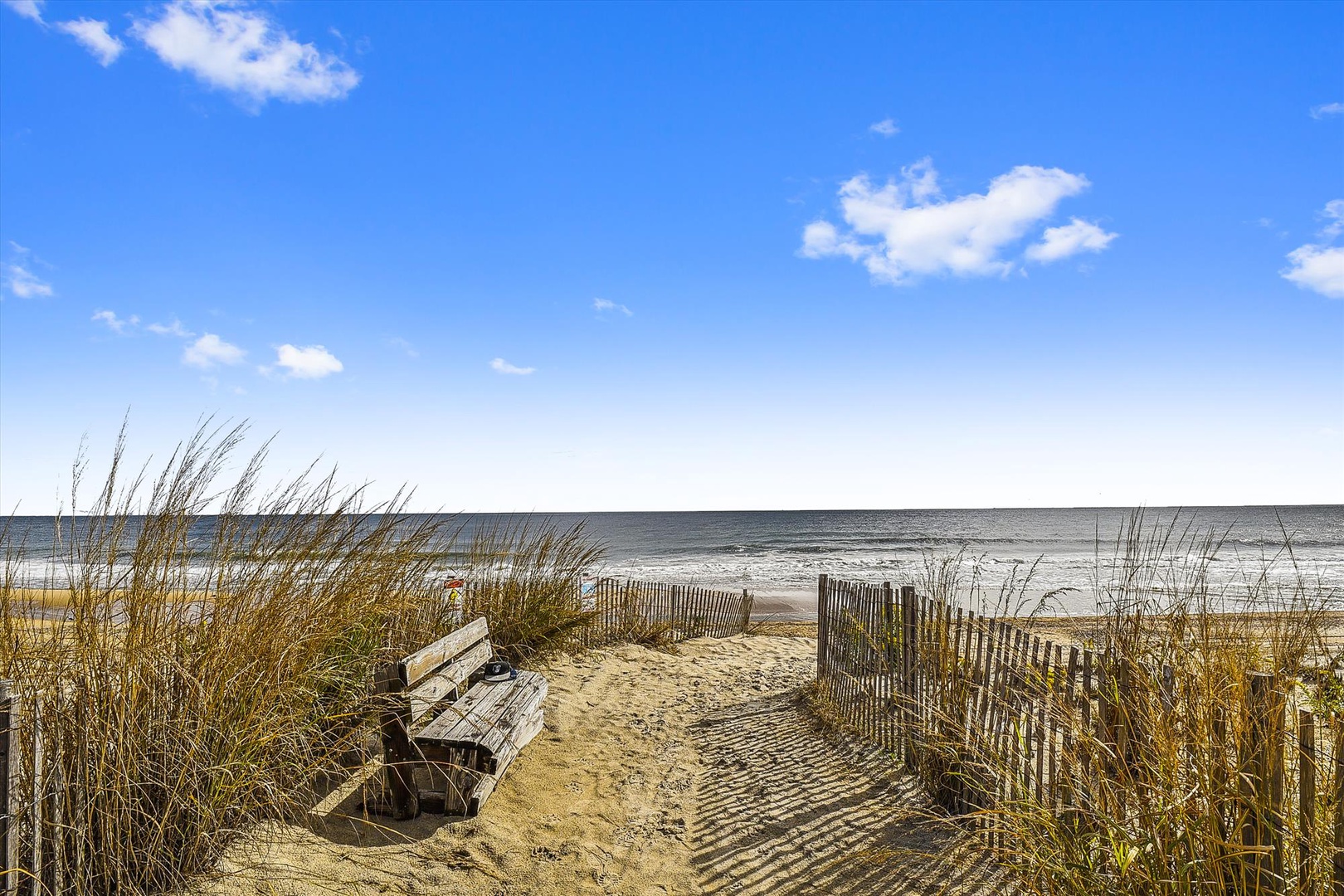 A serene coastal pathway leads through gentle dunes to expansive sandy shores where ocean waves meet horizon under vast blue skies—steps from your beachfront retreat.