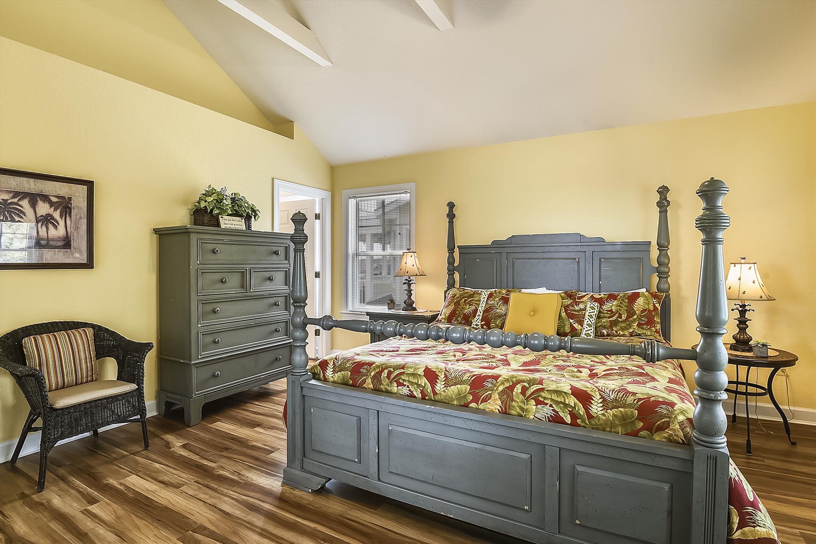Unwind in your bright, cheerful bedroom featuring a stately four-poster bed with tropical-inspired linens, charming vintage furnishings, and hardwood floors that create a warm, inviting retreat.