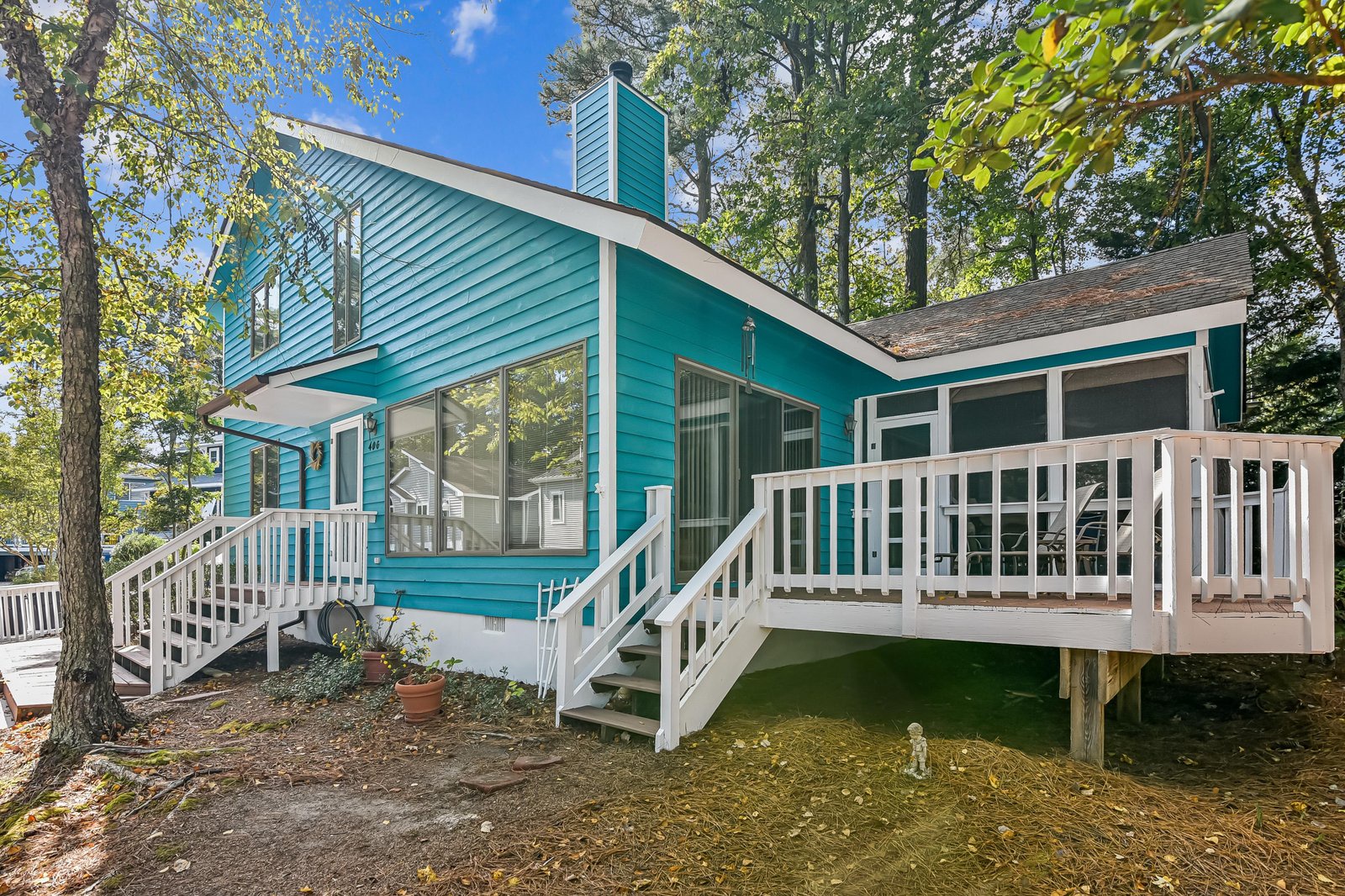 A charming turquoise cottage nestled among mature trees, featuring a spacious wraparound deck and bright, welcoming windows—your peaceful retreat awaits.