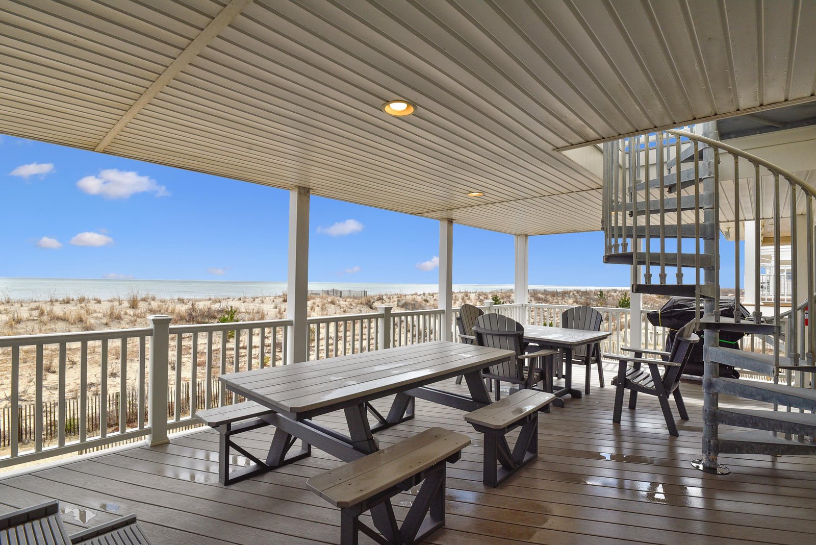 A spacious covered deck with dining and seating areas overlooking a serene beachfront landscape, offering direct access to sandy shores and coastal dunes just beyond.