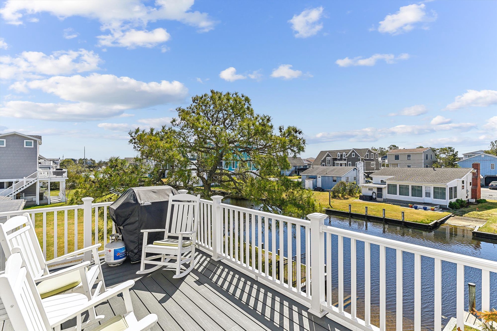 Balcony deck featuring a grill and seating, overlooking a peaceful canal and neighborhood. Part of a coastal residential community with water access and tree-lined views.