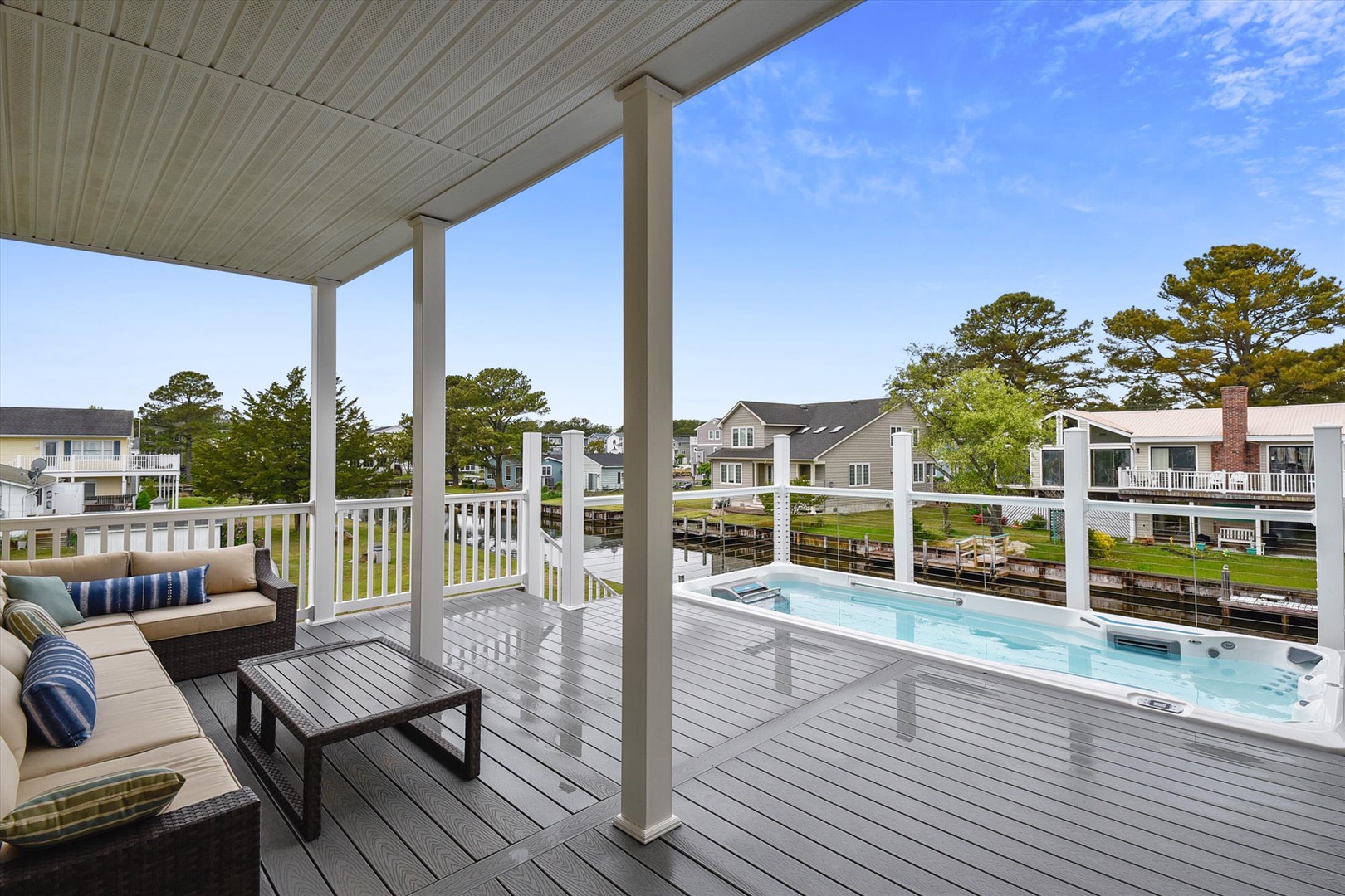 A covered deck overlooks a private hot tub and peaceful canal-side neighborhood, framed by blue skies and mature trees. The property features a waterfront setting ideal for relaxation and scenic enjoyment.