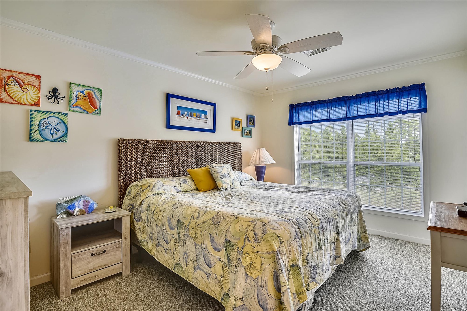 Sink into your plush coastal-themed bedroom where cheerful beach artwork and soft linens invite restful nights under a gentle ceiling fan. Wake up to natural light streaming through large windows overlooking lush greenery.