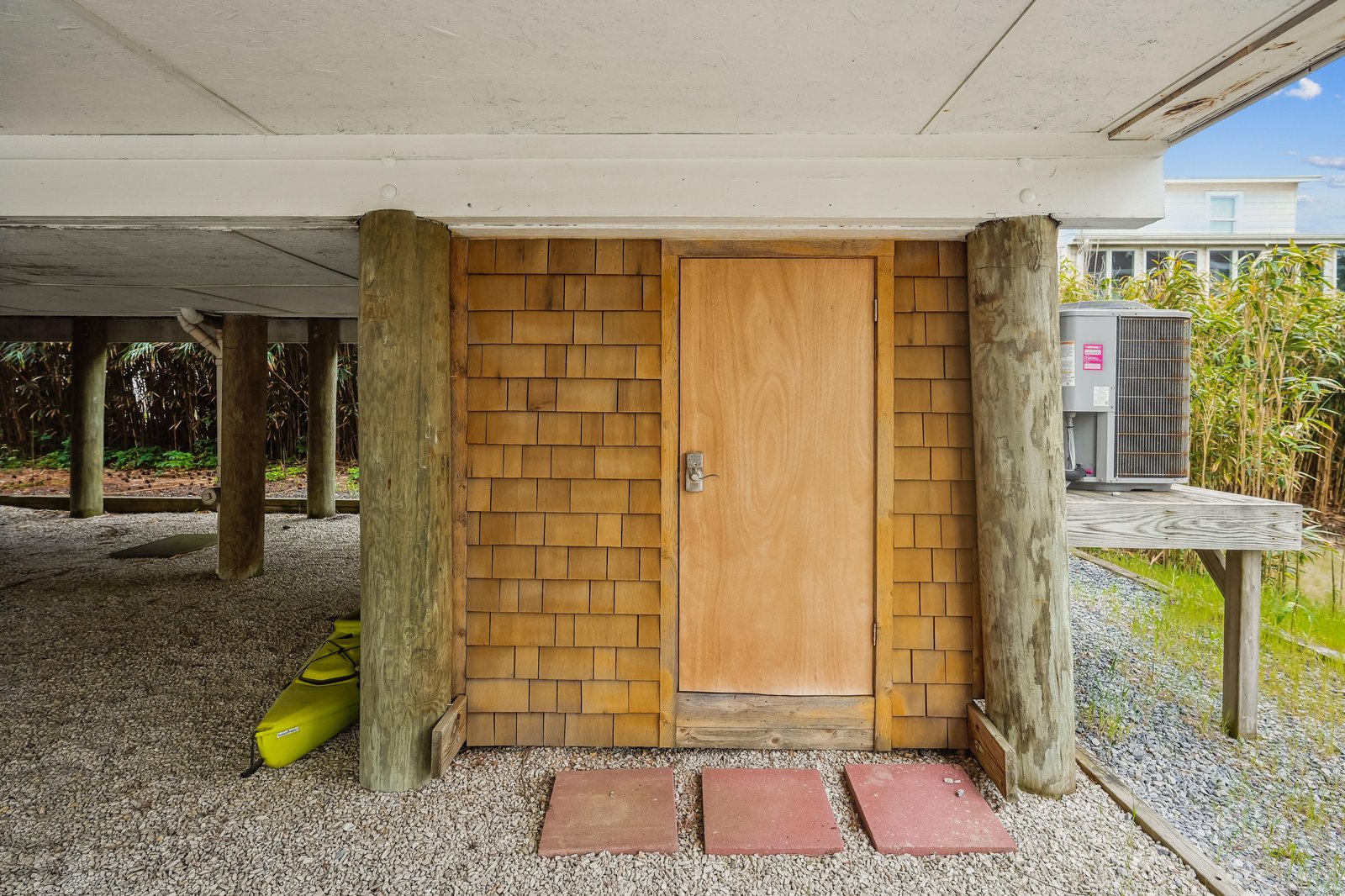 The property's covered ground-level entry showcases coastal-inspired shingle siding and natural wood accents, with parking beneath an elevated structure. A bright yellow kayak rests nearby, hinting at the waterside adventures awaiting you.