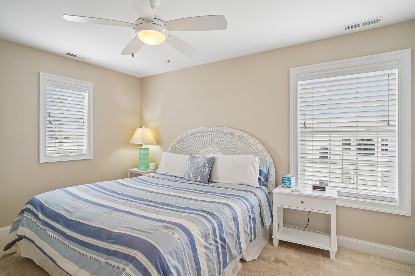 Your serene bedroom awaits with a plush bed dressed in coastal-inspired linens, gentle ceiling fan overhead, and soft natural light filtering through the windows—perfect for restful mornings and peaceful nights.