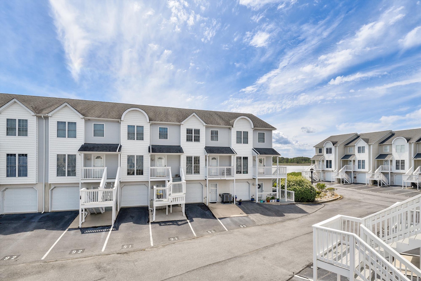 A townhouse community with attached garages, balconies, and parking spaces, situated under a clear blue sky with surrounding greenery.