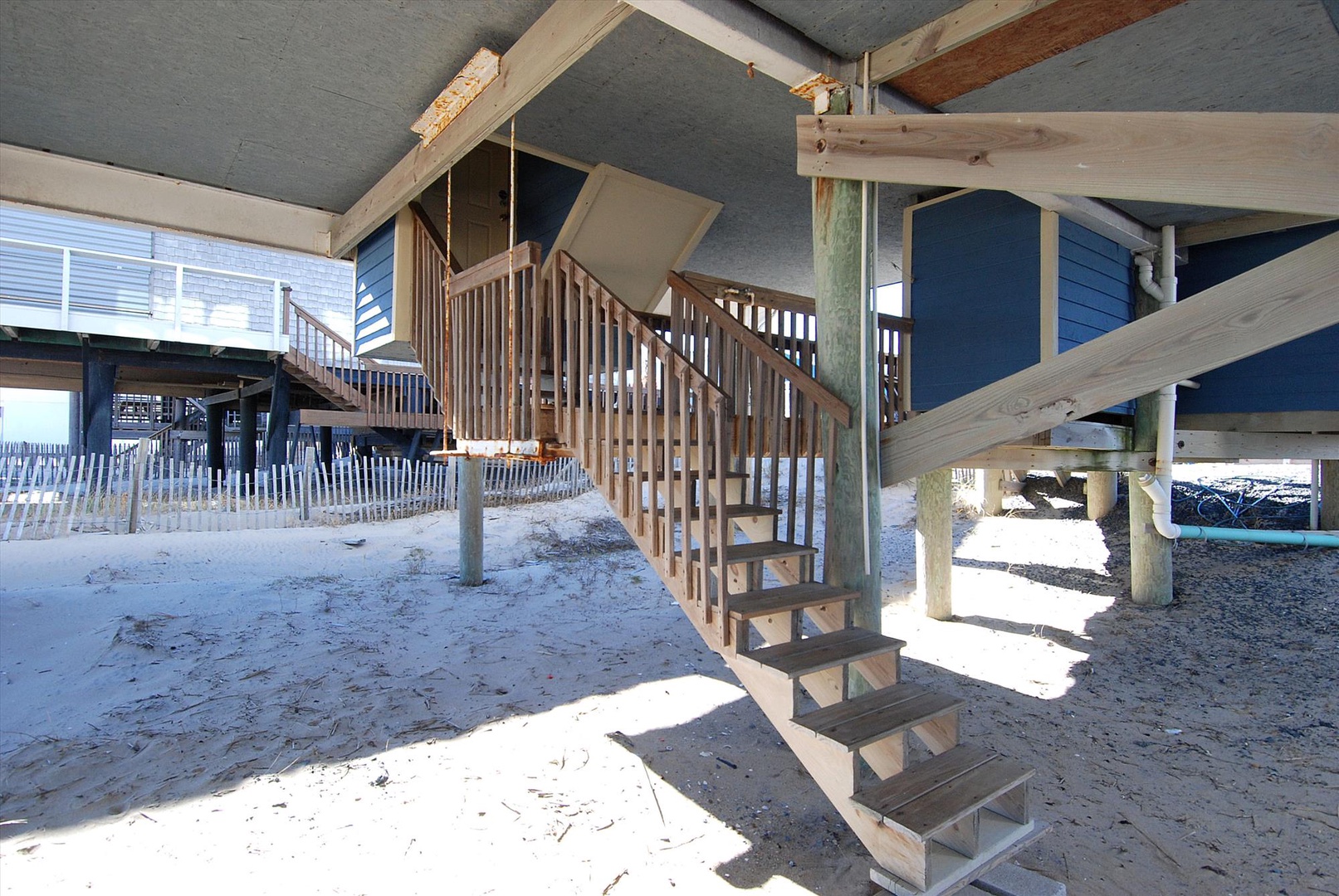 Elevated beach house entrance with wooden stairs leading to blue-sided coastal property on stilts above sandy ground.