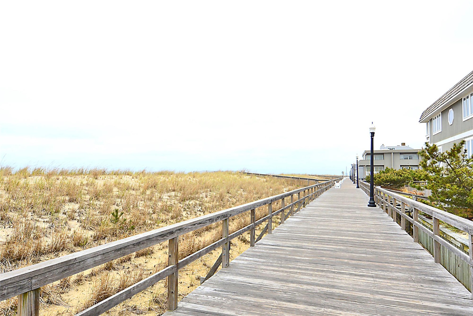 A wooden boardwalk leads through coastal dunes toward the beach, with residential properties visible nearby. The walkway provides convenient access to the sandy shoreline and waterfront area.