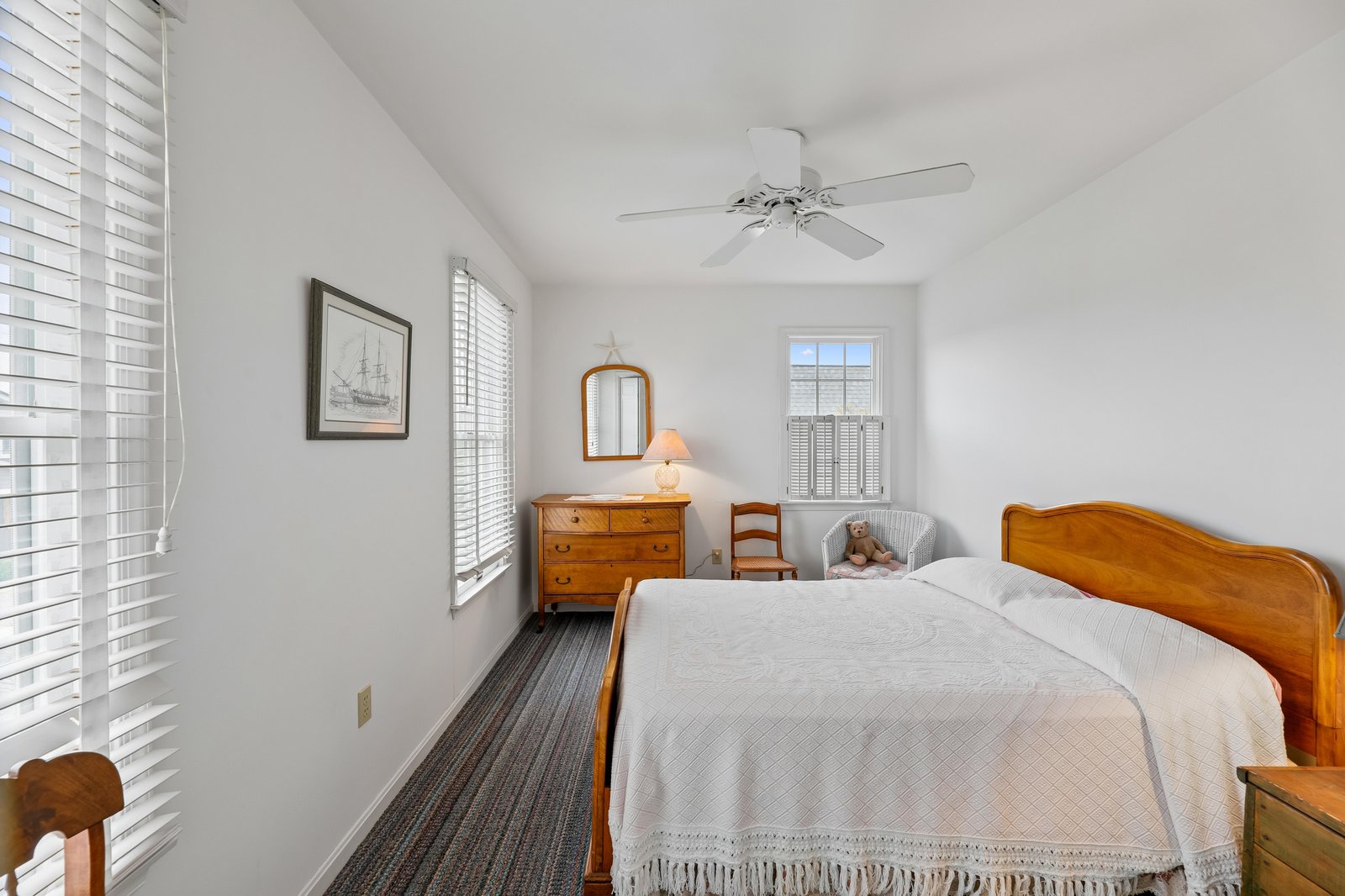 Your peaceful bedroom features warm wood furnishings, crisp white linens, and coastal charm with soft natural light filtering through the blinds. The ceiling fan keeps you comfortable year-round.