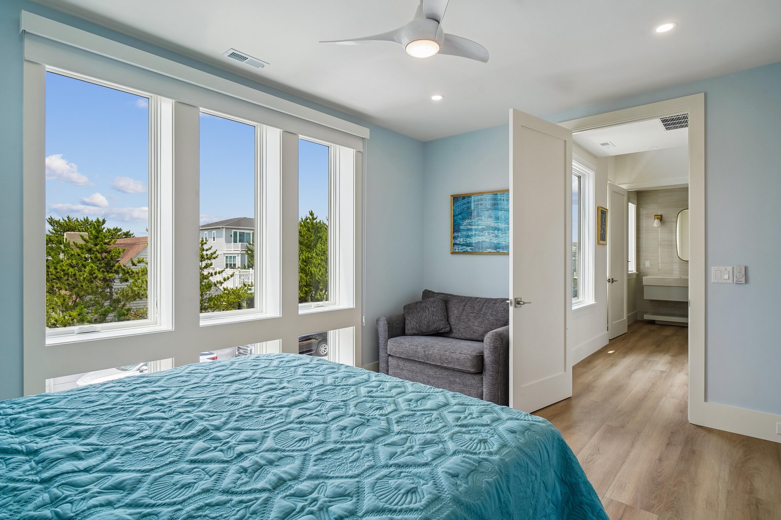 Wake up refreshed in your airy bedroom with coastal vibes, featuring soft blue tones, bright windows framing neighborhood greenery, and a cozy loveseat perfect for your morning coffee.