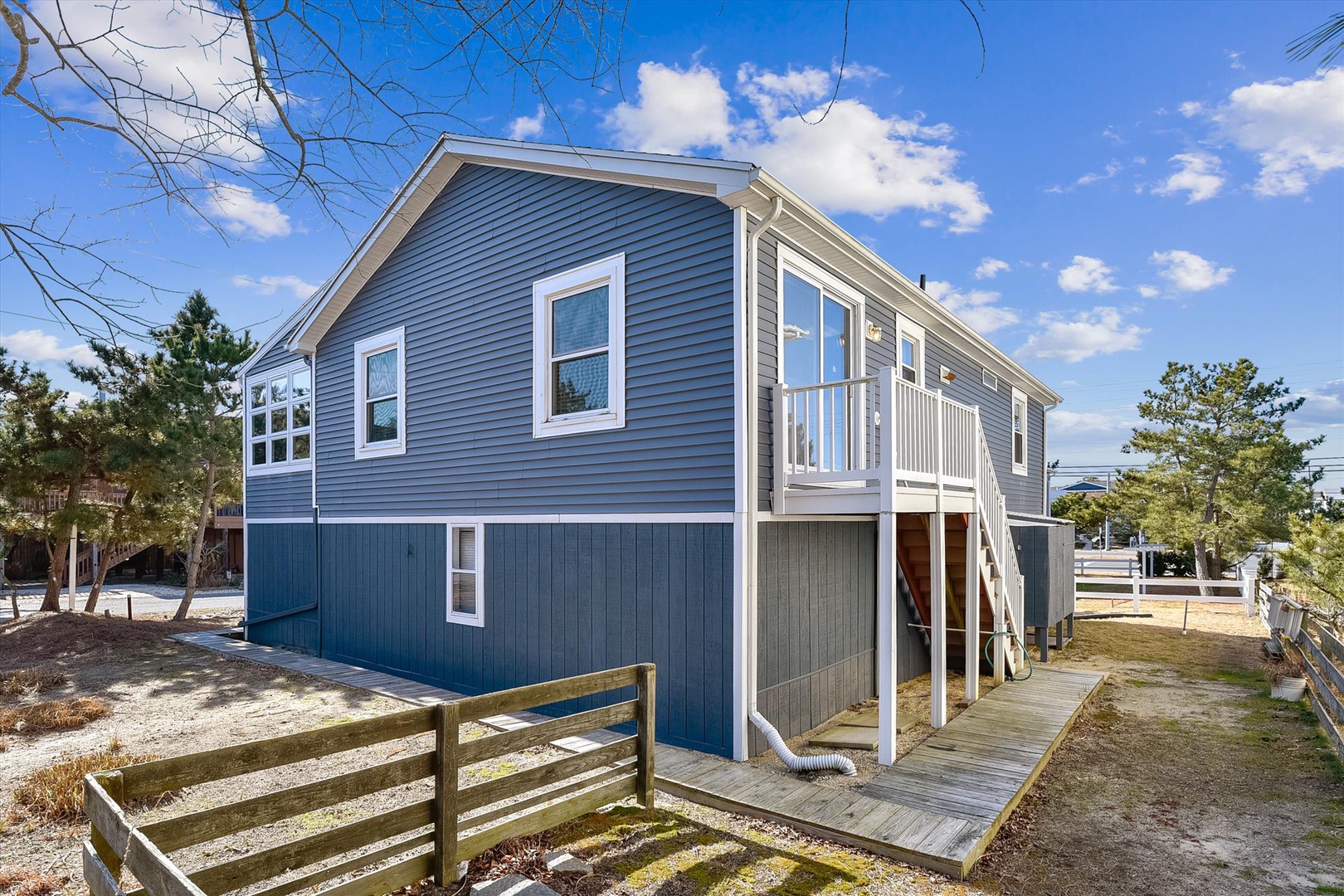 A charming coastal rental featuring modern blue siding with white trim, surrounded by natural landscaping. The elevated design with wooden stairs provides easy access to the main entrance on a bright, sunny day.