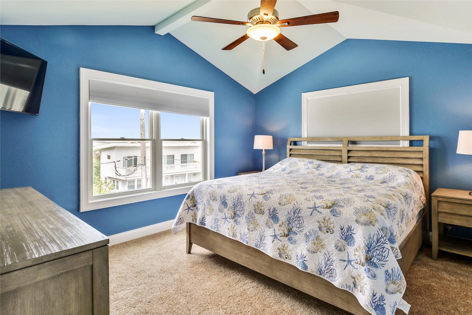 Drift off in this tranquil coastal bedroom with soothing blue walls, a plush queen bed dressed in ocean-themed linens, and vaulted ceilings with a gentle ceiling fan to keep you comfortable year-round.