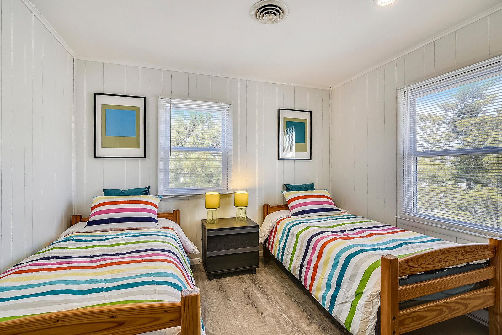 Step into this cheerful bedroom with twin beds dressed in playful rainbow stripes. Bright windows frame leafy views, and the crisp white paneling keeps the space feeling fresh and airy—perfect for your vacation mornings.