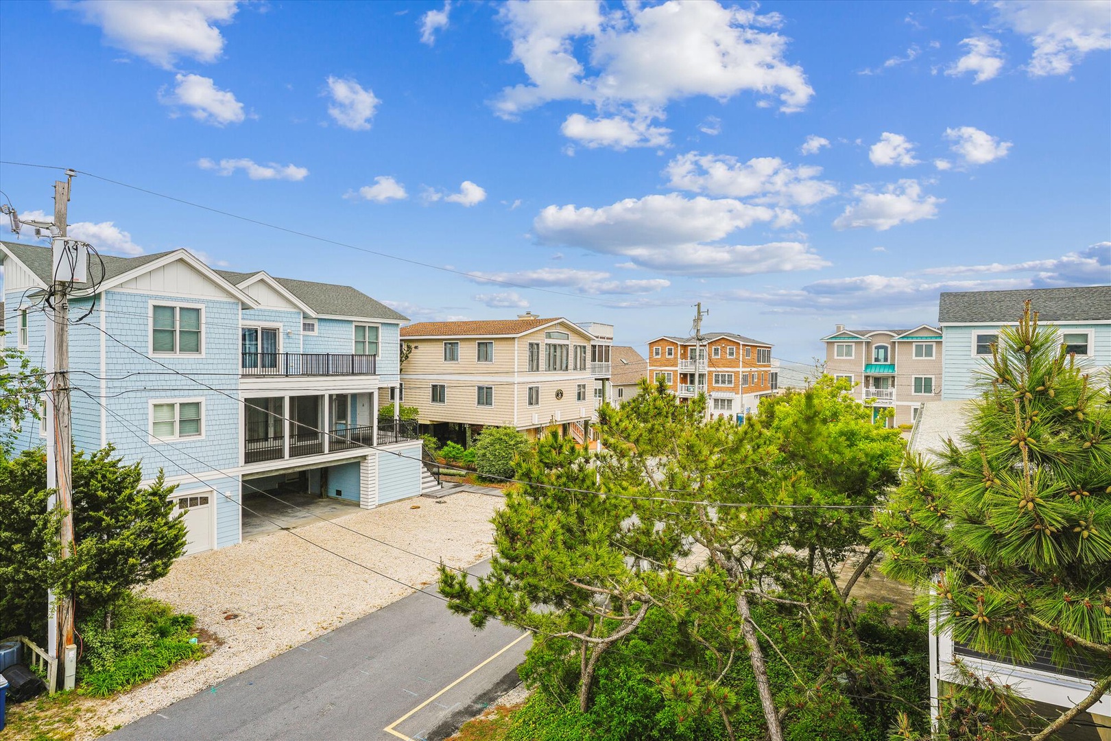 A coastal residential neighborhood with vacation homes, featuring the property's exterior surrounded by lush greenery and neighboring beach houses under a bright blue sky.