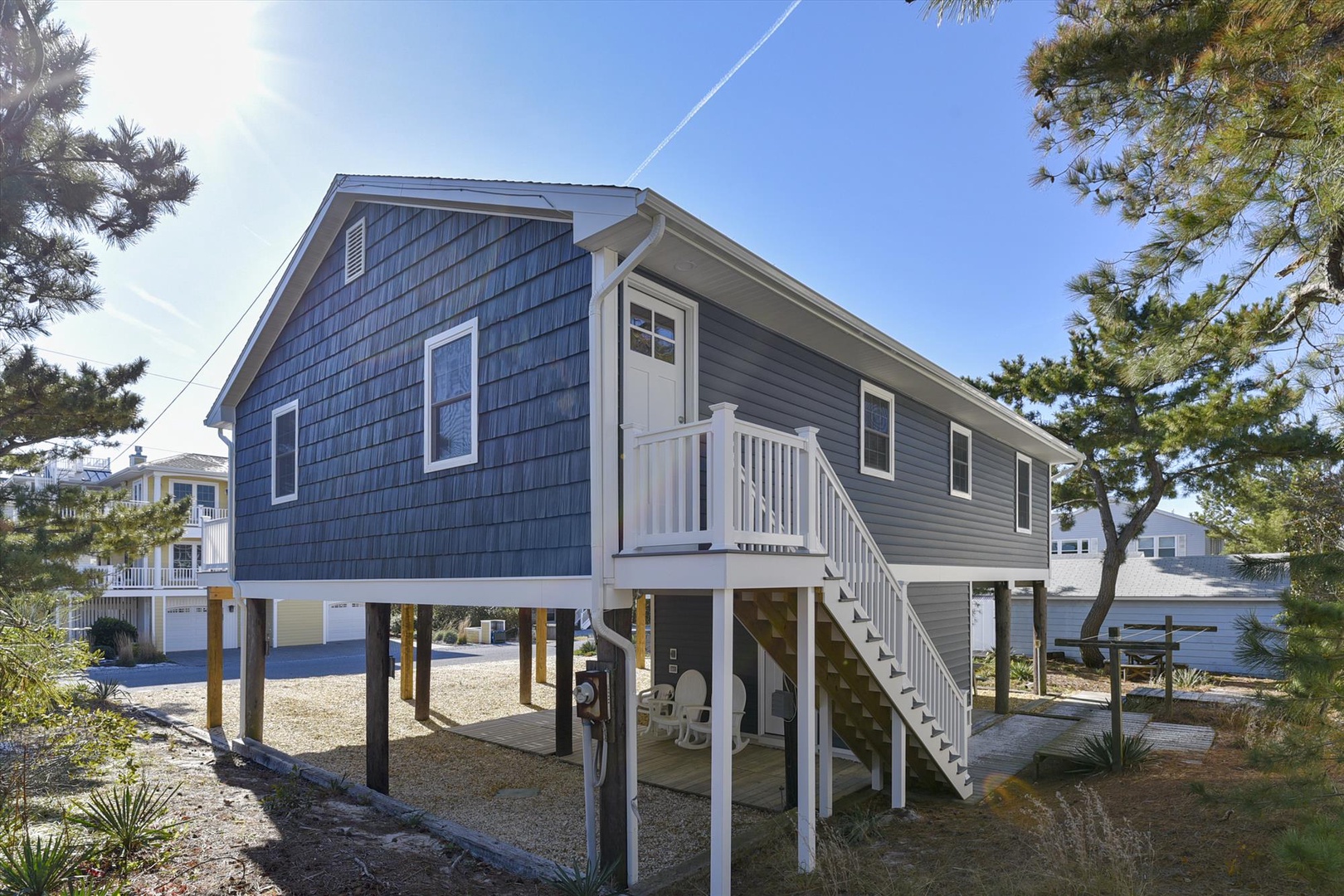 Charming elevated coastal cottage with blue shingled siding nestled among mature trees in a peaceful waterfront neighborhood.