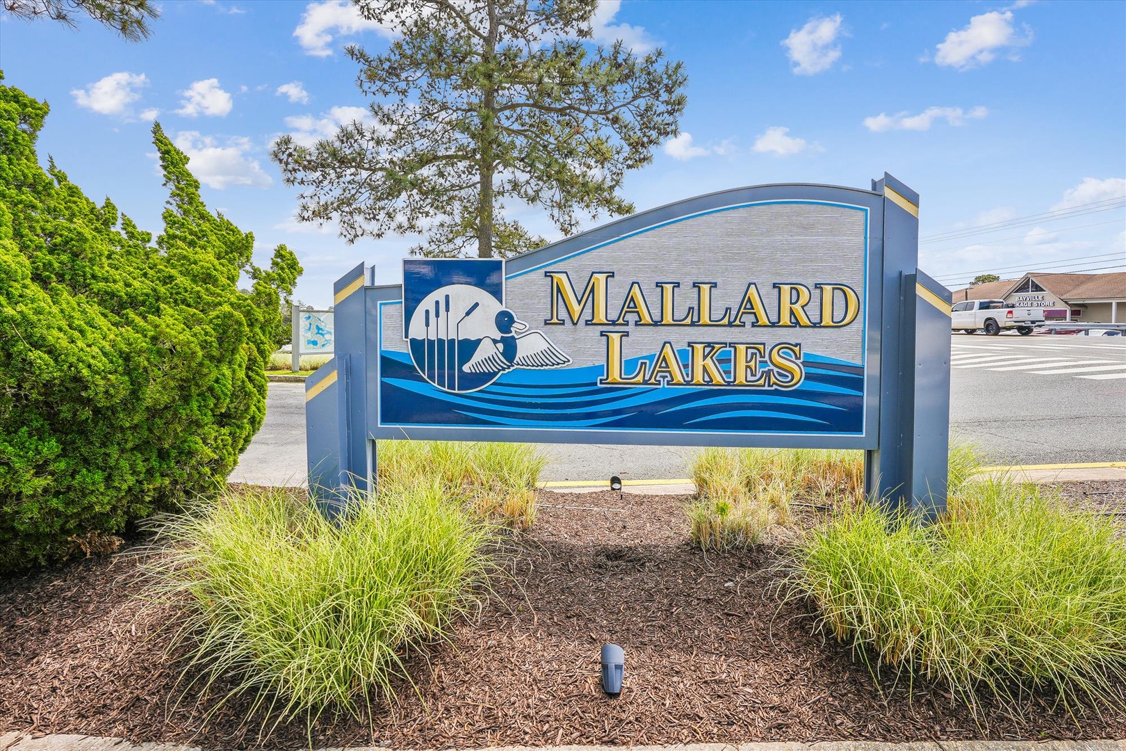 The entrance sign at Mallard Lakes marks the property location. The blue and gold design features a duck motif, set in landscaped surroundings near the parking area.