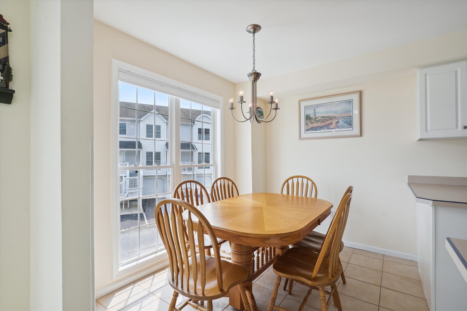 Your sun-drenched dining space features a warm wood table for six beneath an elegant chandelier, with coastal artwork completing the inviting atmosphere—perfect for shared meals and memories.