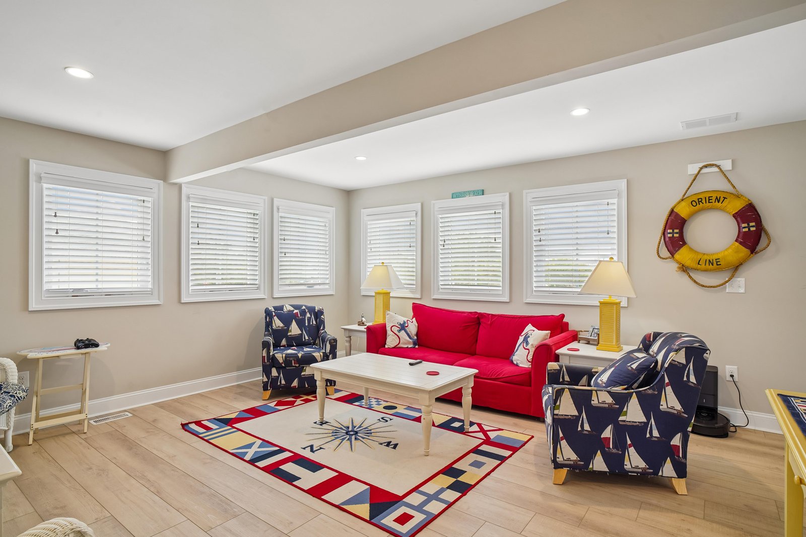 Settle into the charming nautical-themed living room with bright red sofas and sailboat armchairs. Sunlight streams through white blinds, illuminating the compass rug and cheerful yellow accents that make this space cozy and spirited.