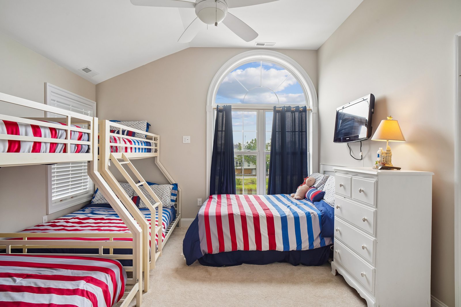 Your cheerful bedroom awaits with cozy bunk beds and bright red-and-blue striped bedding. A ceiling fan keeps you cool, and large windows invite natural light into your comfortable retreat.