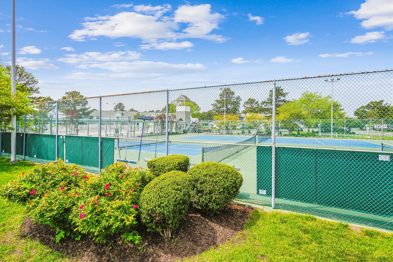 Well-maintained tennis courts within a landscaped resort area, framed by manicured hedges and colorful seasonal flowers under a clear blue sky.