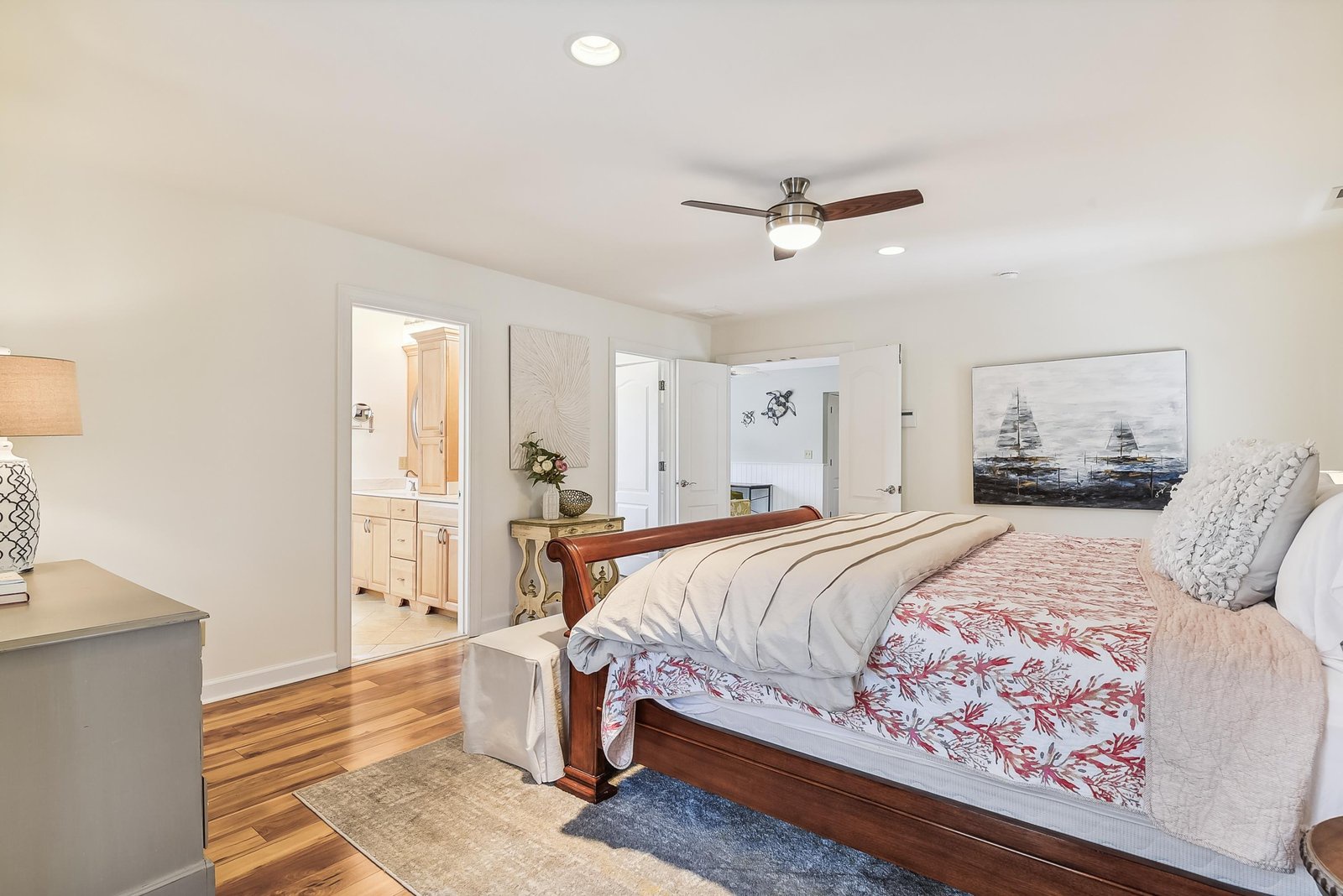 Your serene bedroom retreat features warm hardwood floors, a plush bed with coastal-inspired linens, and a gentle ceiling fan overhead. Through the open door, you'll glimpse your private ensuite waiting to pamper you after each beach day.
