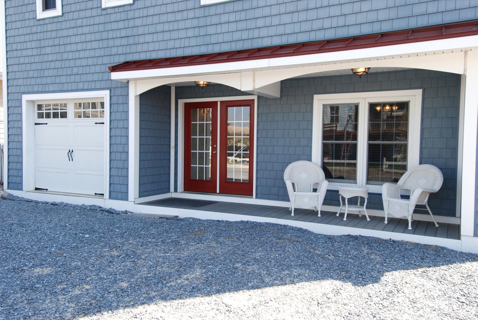 The property features a charming covered porch with welcoming seating and a red double-door entryway. Classic blue shingle siding and convenient garage access complete the inviting exterior.