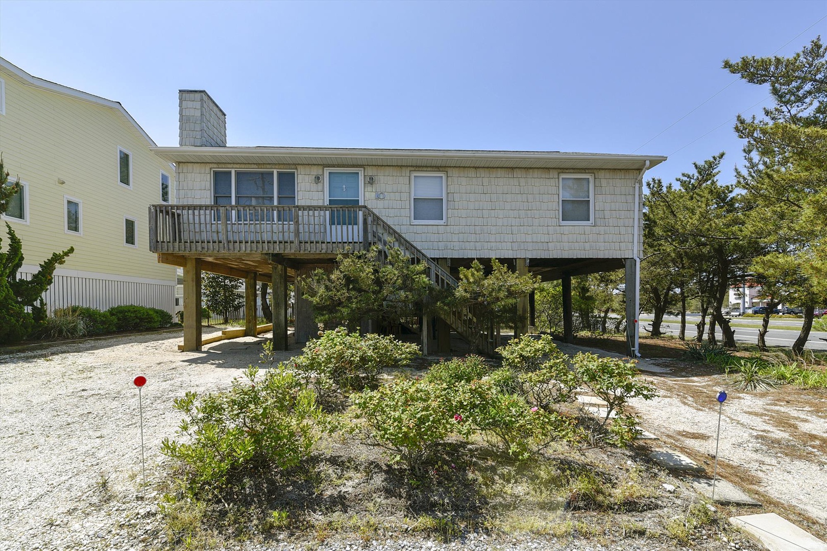 Coastal beach house elevated on stilts with private deck and landscaped grounds. The property features covered parking below and natural greenery surrounding the home.