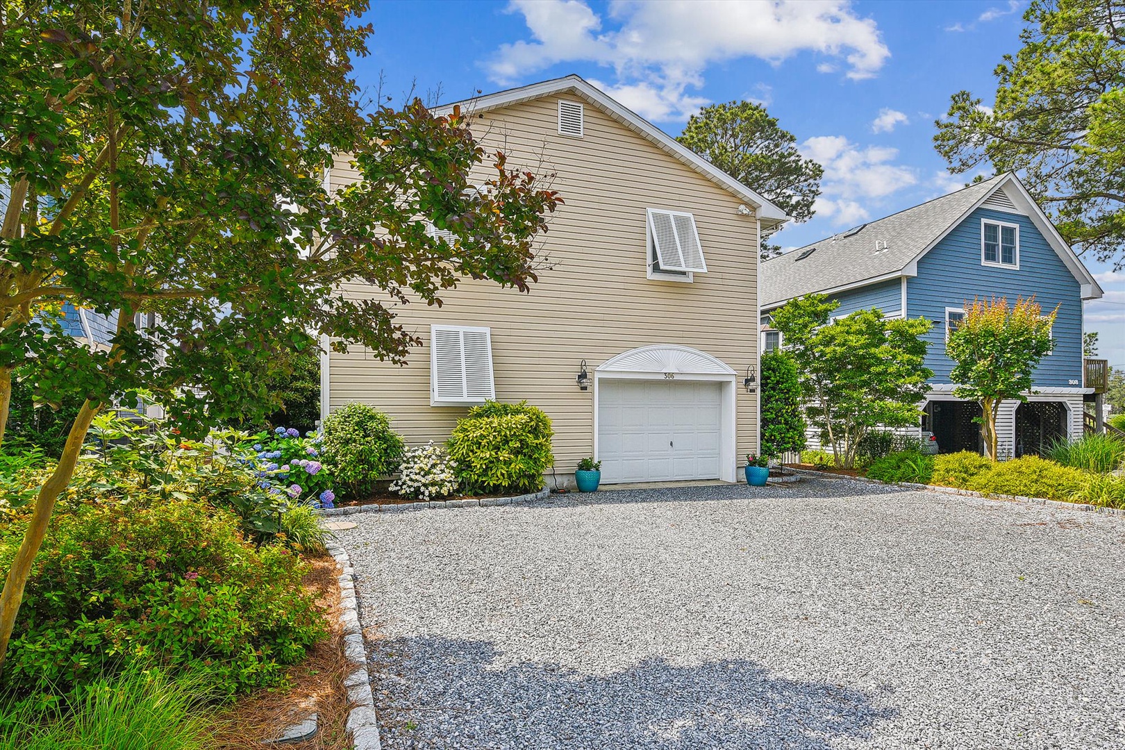 A charming coastal home with gravel driveway nestled in a quiet residential neighborhood, featuring manicured gardens and mature trees surrounding the property.
