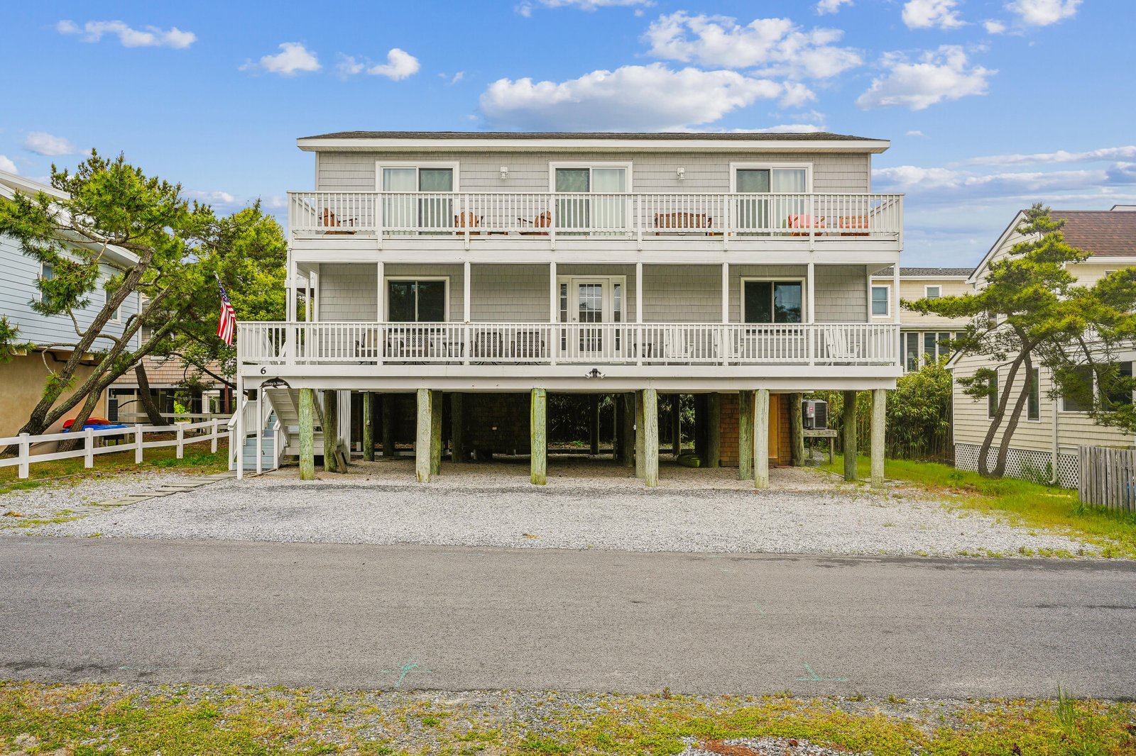 A spacious coastal retreat elevated on stilts, featuring multiple balconies and generous parking beneath the home, set in a peaceful beach neighborhood with mature trees framing the property.