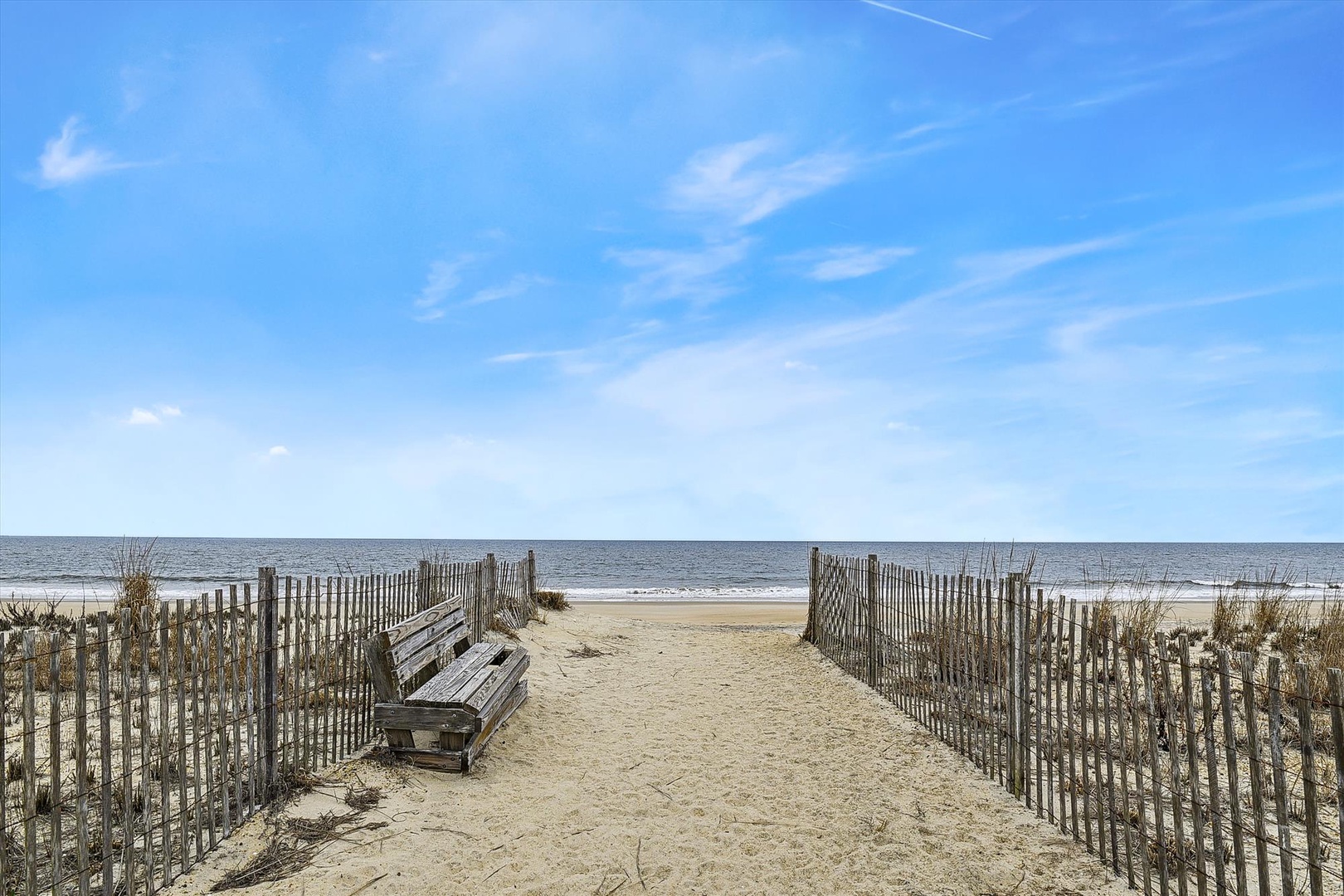 A tranquil beach access pathway leads directly to the sandy shore, with wooden fencing guiding the approach and a thoughtfully placed bench for soaking in the coastal serenity.