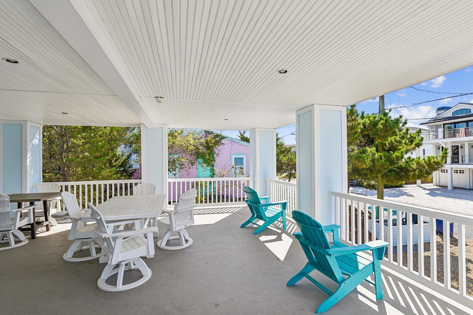 A spacious covered porch features turquoise Adirondack chairs and a white dining set, offering a bright, coastal retreat with neighborhood views.
