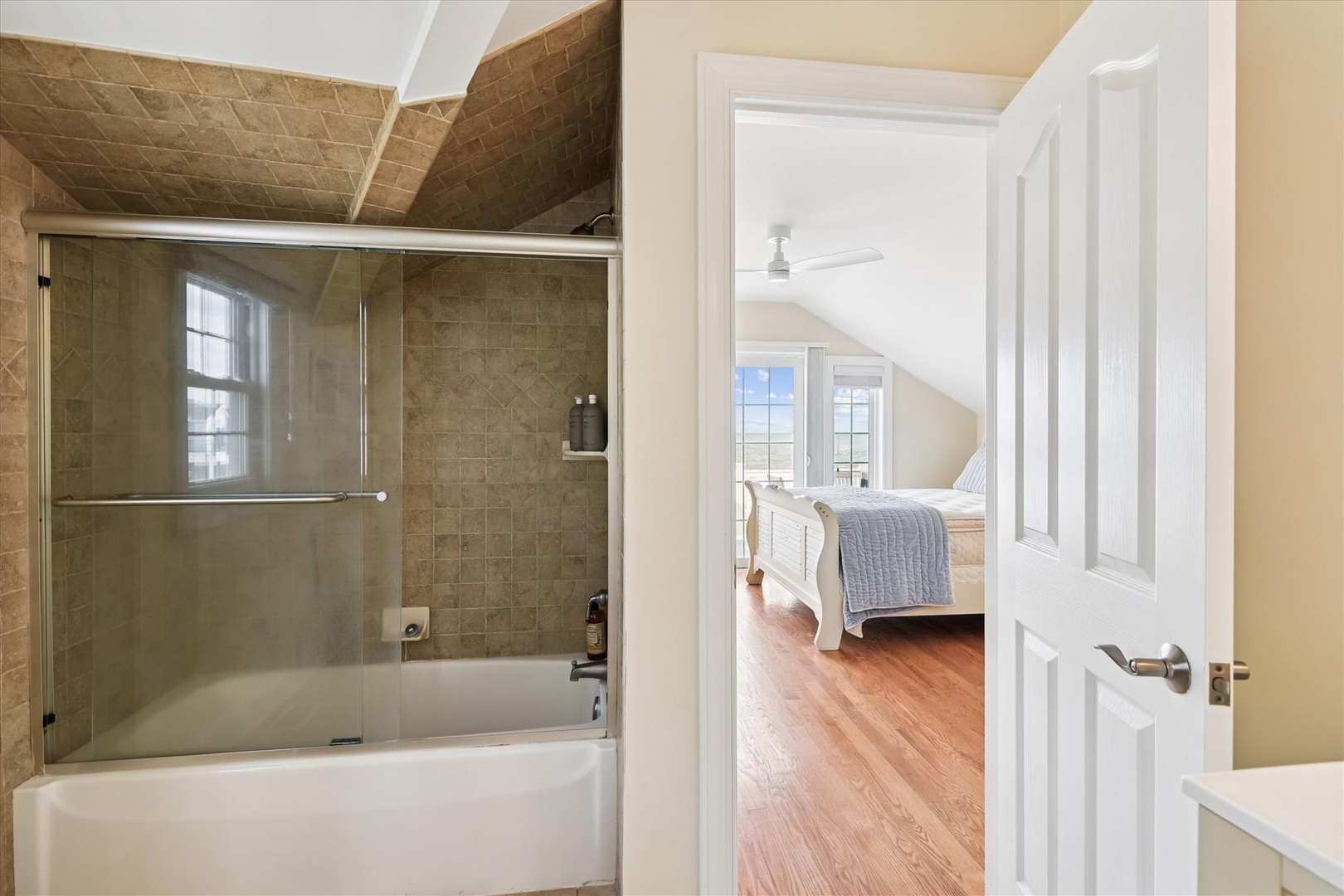 Step into your private ensuite with a sleek tub-shower combo and warm stone tile, connected to your serene bedroom retreat with bright windows and hardwood floors.