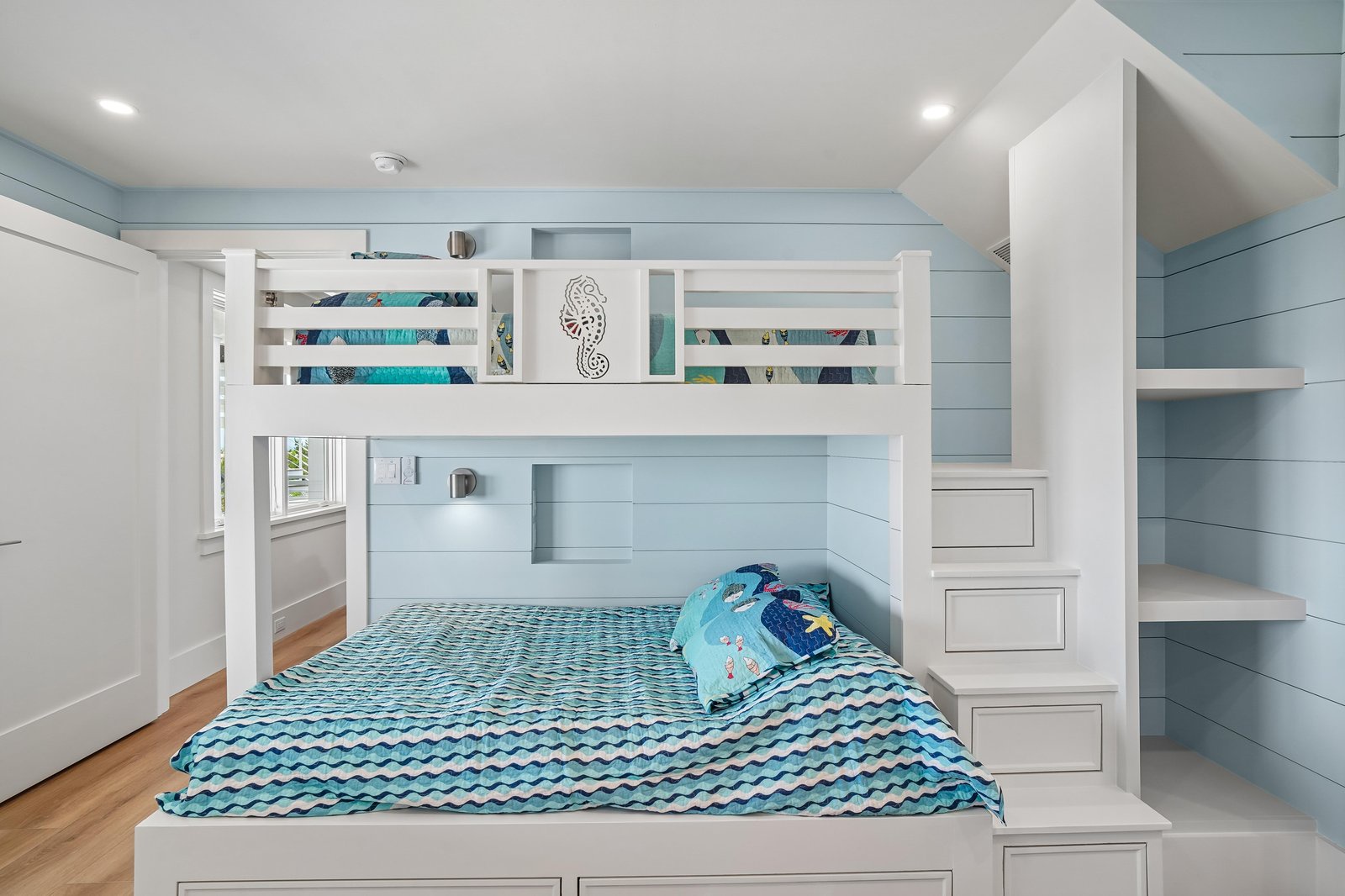 This playful bedroom features custom bunk beds with built-in stairs and storage—perfect for kids who love to explore and make memories. The coastal blue walls and bright natural light create a cheerful, inviting retreat.
