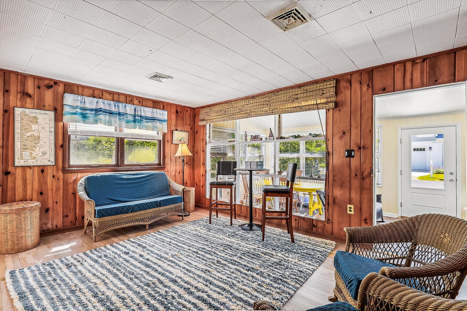 Step into your breezy coastal retreat with wicker seating, warm wood walls, and bright windows framing views of greenery and open sky.